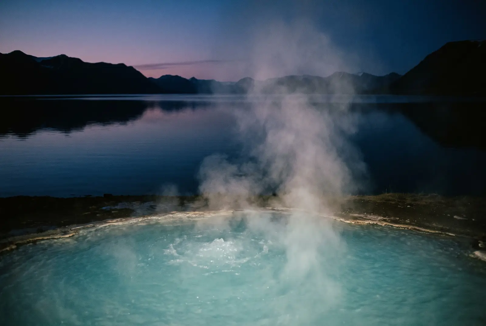 Steam rising from a turquoise geothermal pool beside a mirror-calm Eastfjords lake